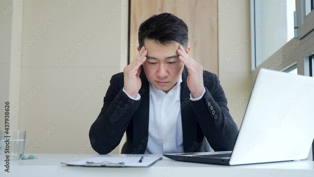 young asian man, office worker sitting at workplace holding hands massages forehead and head with severe pain. Male at work with a headache. Up close face of sufferer is overloaded with sick. indoors young asian man, office worker sitting at workplace holding hands massages forehead and head with severe pain. Male at work with a headache. Up close face of sufferer is overloaded with sick. indoors