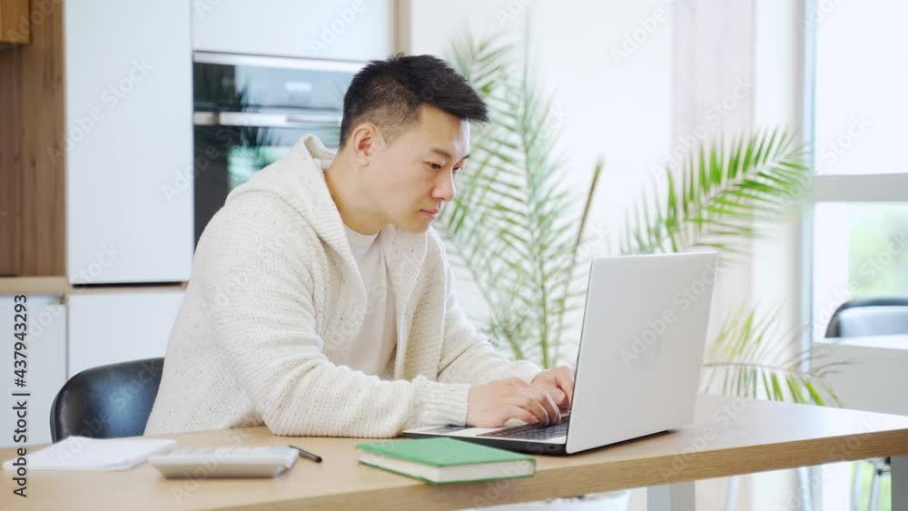 young asian man sitting at home a table and working at a computer or laptop. Male student learns online. E-learning to write a notebook. Internet lessons distance. Remote Education. guy studying young asian man sitting at home a table and working at a computer or laptop. Male student learns online. E-learning to write a notebook. Internet lessons distance. Remote Education. guy studying