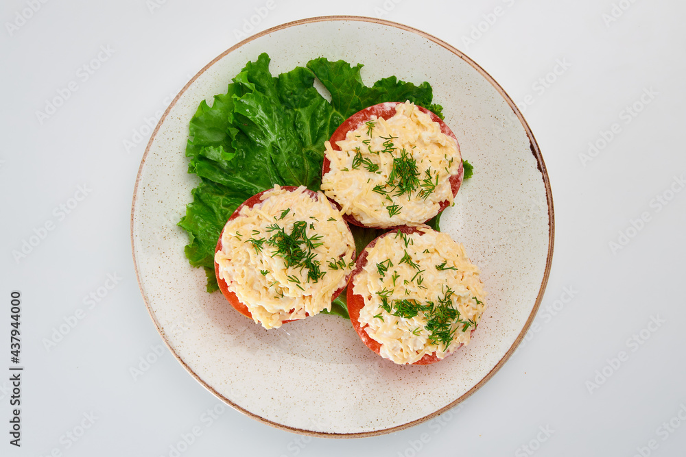 Three halves of fresh tomato with grated cheese, garlic, white sauce, sprinkled with finely chopped fresh dill, with green lettuce leaves in a white round plate on a white background. Three halves of fresh tomato with grated cheese, garlic, white sauce, sprinkled with finely chopped fresh dill, with green lettuce leaves in a white round plate on a white background.