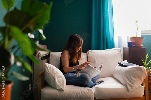 Young red-haired woman, reading a book in a couch surrounded by plants. Leisure indoors entertainment.