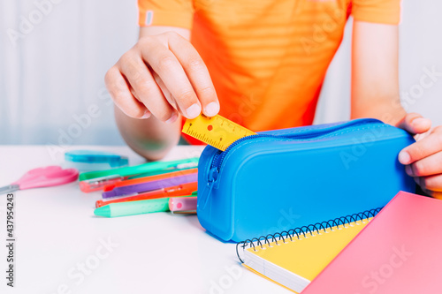 Boy keeping inside his blue pencil case all a small ruler and pencils, pencils, scissors, etc. Colorful notebooks on white table. Back to school concept photo
