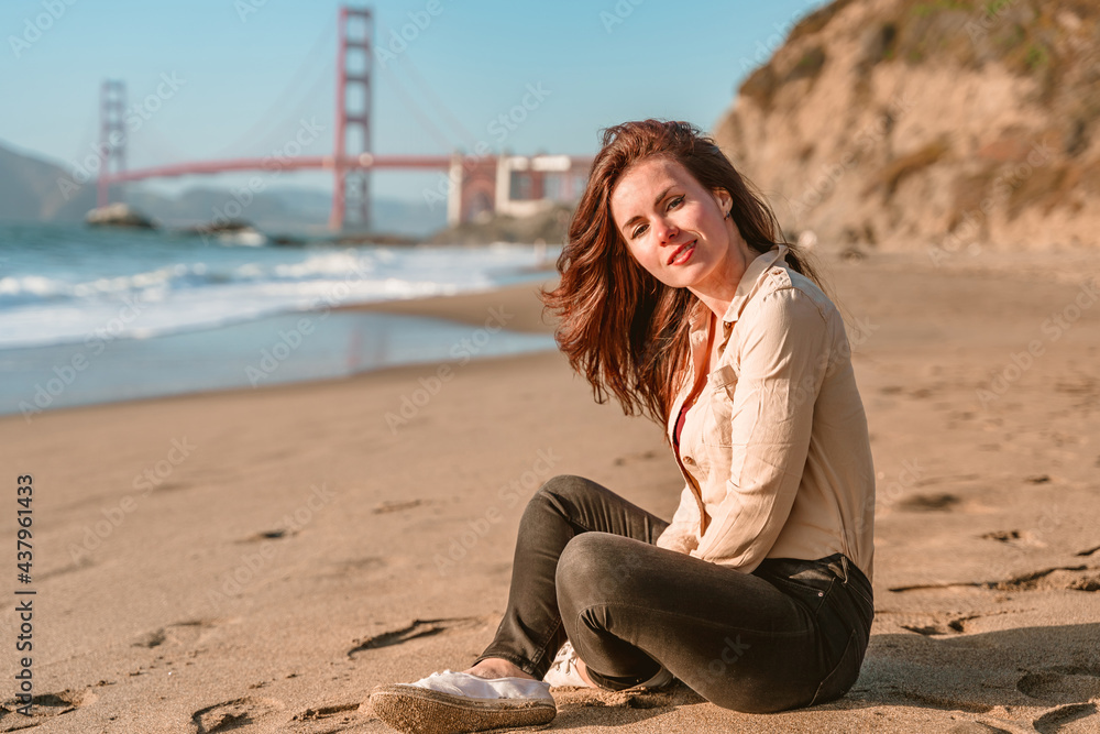 Cute young woman sitting on the sand on the beach overlooking the Golden Gate Bridge in San Francisco