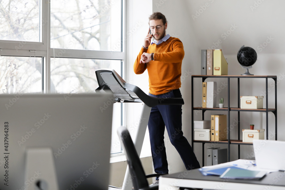 Fototapeta premium Young man talking by mobile phone while running on treadmill in office
