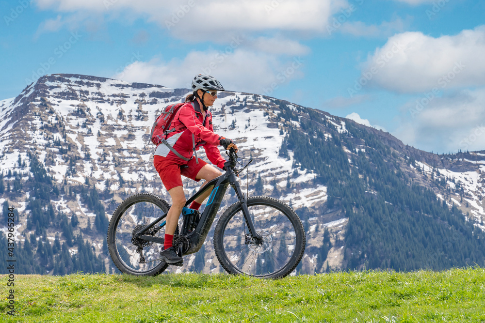 smiling senior woman riding her electric mountain bike on a sunny day in early spring with yello flowers on the meadows below the snow capped mountains of Nagelfluh chain near Oberstaufen, Allgaeu 