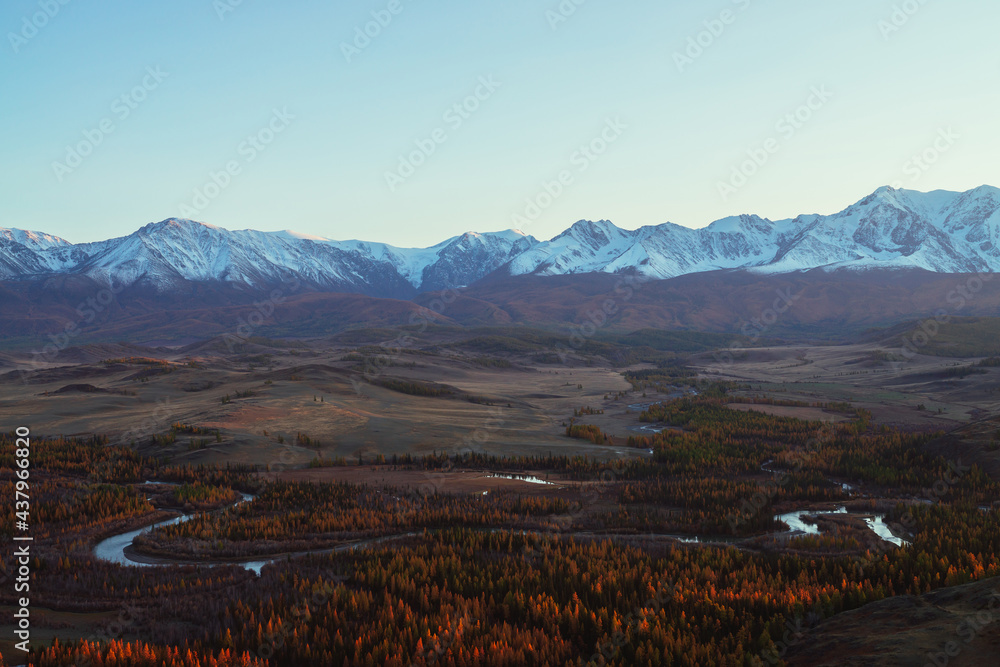 Awesome autumn landscape with mountain river in forest valley and great snow-covered mountain range in red sunset sunshine. Spectacular view from hill to snowy mountains and autumn valley in sunlight.