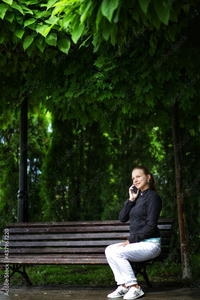 a blonde in white pants and a black jacket sits on a bench under a lush green catalpa, talks on the phone and smiles a blonde in white pants and a black jacket sits on a bench under a lush green catalpa, talks on the phone and smiles