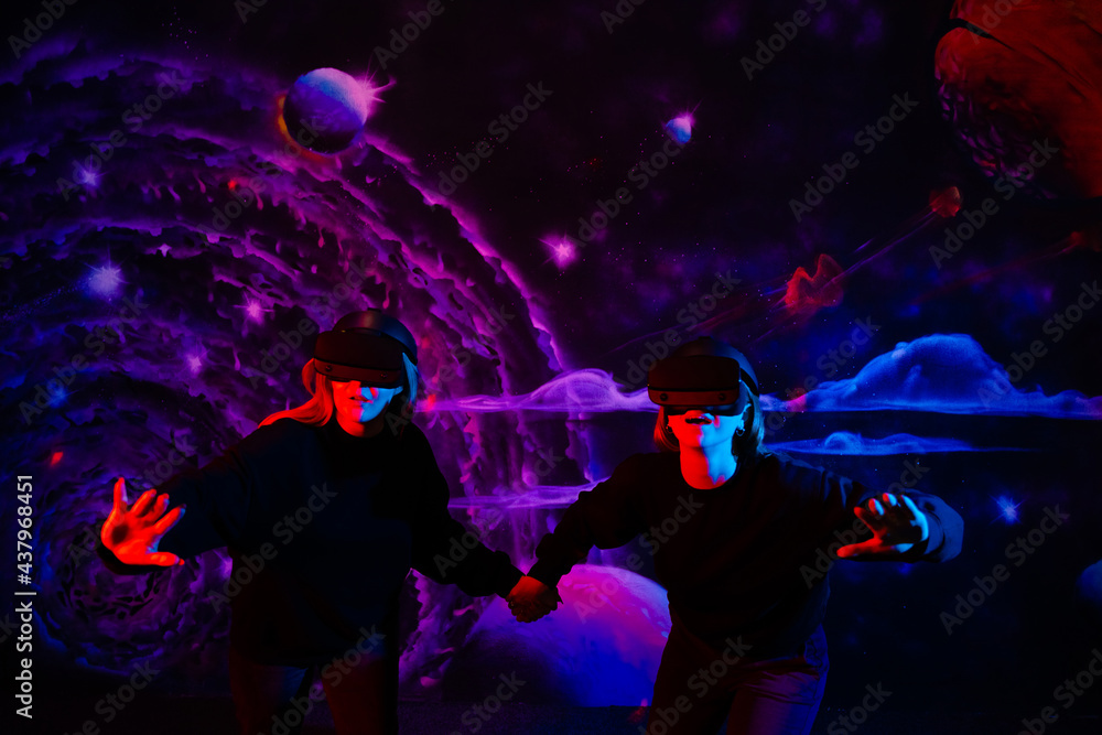 Two girls friends in virtual glasses are holding hands in the playroom in neon light