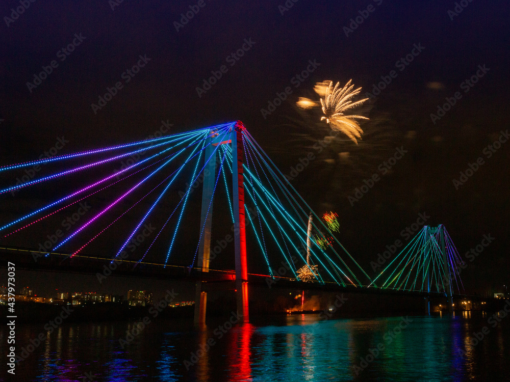 Obraz premium Blurred motion of bright flashes of fireworks in the night sky above the bridge. Fireworks in honor of Victory Day in Krasnoyarsk, Russia.