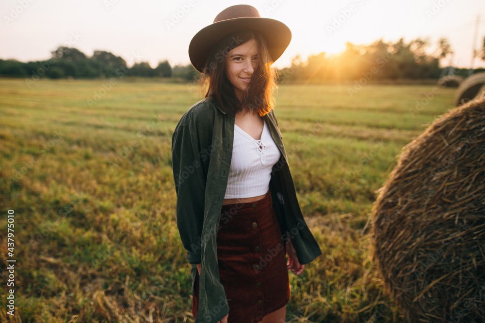 Obraz premium Beautiful stylish woman in hat walking at haystacks in sunset light in summer field. Atmospheric tranquil moment in countryside. Young female enjoying evening at hay bale in warm sunshine