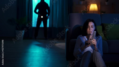 terrified young woman sitting on the floor with a gun in her hands and sees a robber with a fla in her house