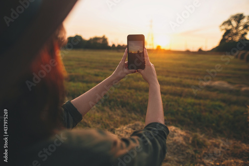 Stylish woman taking photo of sunset on phone in summer field. Young female holding smartphone and capturing evening warm sunshine in countryside. Atmospheric beautiful moment