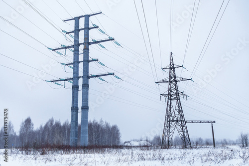 High voltage power line towers in a winter climate. It can be seen snowing. Snowy field and forest line on background