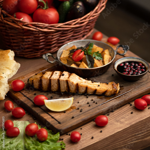 Luxury seafood lunch. Grilled red fish with vegetables and pomegranate sauce on wooden cutting board. Basket with tomatoes and grilled salmon steak and lemon on table. Soft focus.