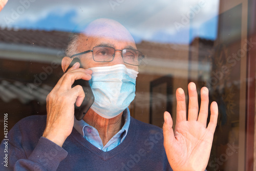 Portrait od an old senior man with glasses and medical face mask talking on the phone behind a glass with reflection of buildings and sky, hand on the glass. Scene of COVID or Coronavirus pandemic.