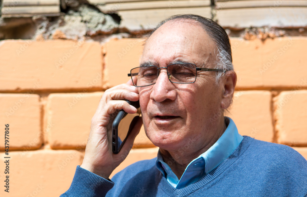 Portrait od an old senior man with glasses and talking on the phone outside with a orange brick wall in the background. Scene of COVID or Coronavirus pandemic. Portrait od an old senior man with glasses and talking on the phone outside with a orange brick wall in the background. Scene of COVID or Coronavirus pandemic.