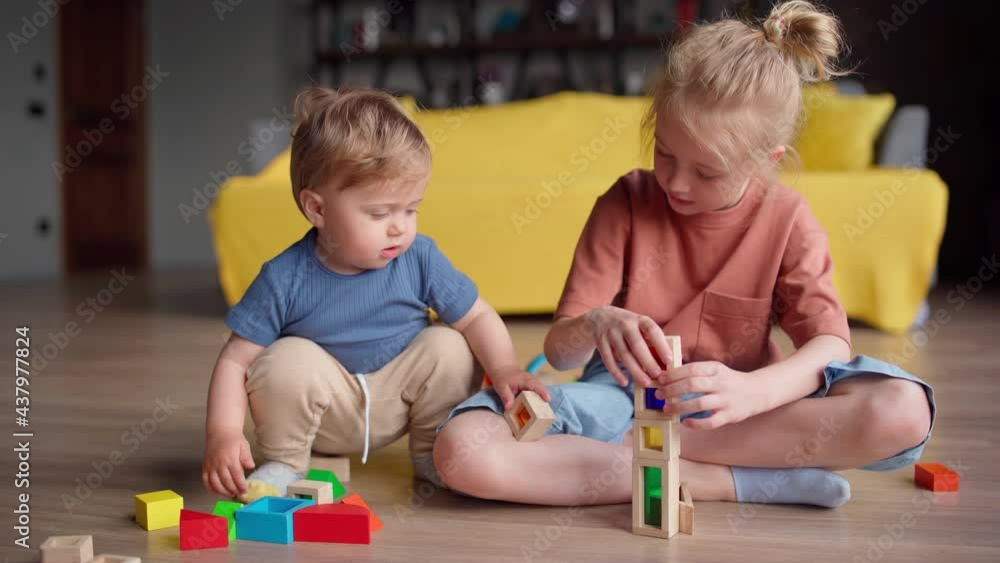 Little girl and baby boy playing construction set sitting on floor at home. Toddler brother trying to help sister with building toy but breaking construction Little girl and baby boy playing construction set sitting on floor at home. Toddler brother trying to help sister with building toy but breaking construction