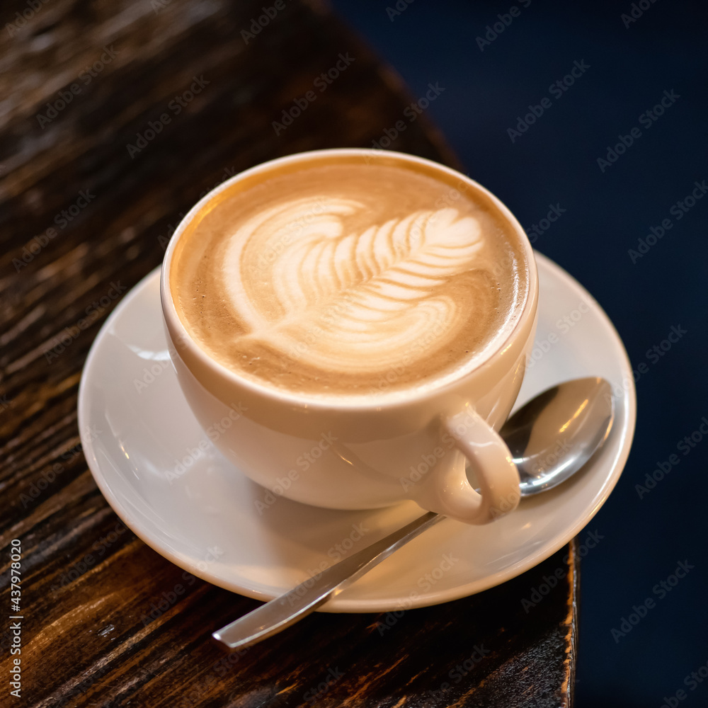 Coffee with milk. Cappuccino with creamy foam. Rest break from business with natural energy drink. Foam drawing. Cup of aromatic coffee with spoon and saucer on a dark background. Coffee with milk. Cappuccino with creamy foam. Rest break from business with natural energy drink. Foam drawing. Cup of aromatic coffee with spoon and saucer on a dark background.