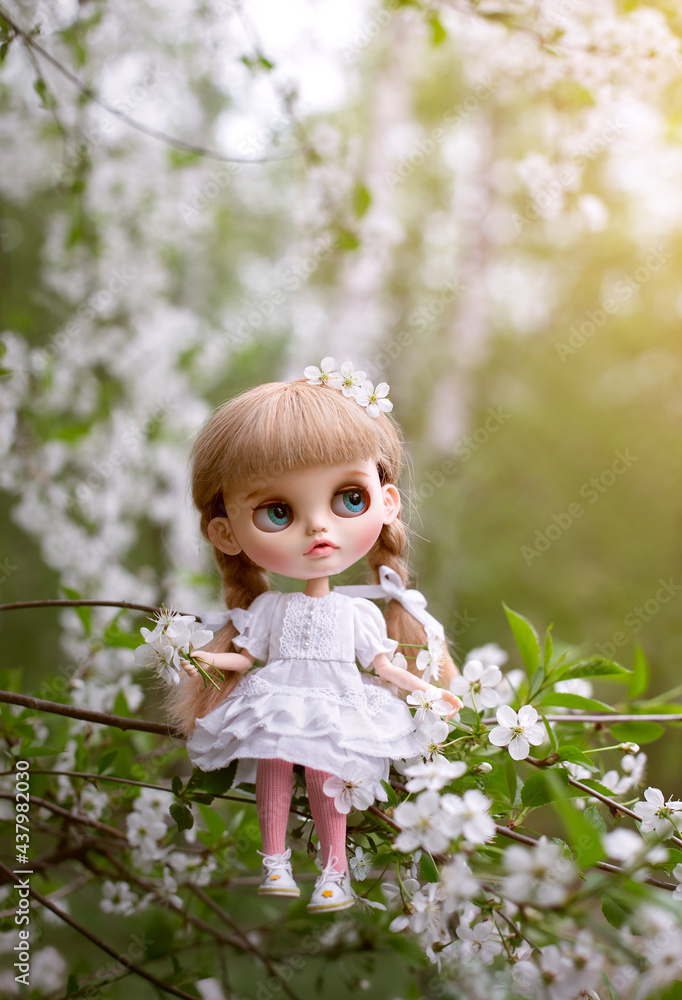 Blonde doll blythe with bangs in a white dress sits on a twig in a spring cherry orchard among white flowers
