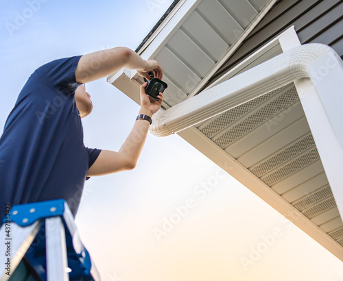 Technician on ladder installing and adjusting wireless security video camera underneath a roof for private house, monitor and recorder. Set up outdoor surveillance system. Safety and privacy concept.