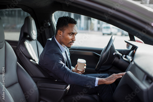 Side view of focused african man sitting on driver's seat with cup of coffee and using dashboard for navigation. Businessman in formal wear taking stop for checking direction. © sofiko14
