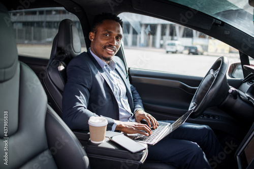 African american man in business suit sitting on driver's seat of modern electric car and typing in wireless laptop. Busy person with portable computer. © sofiko14