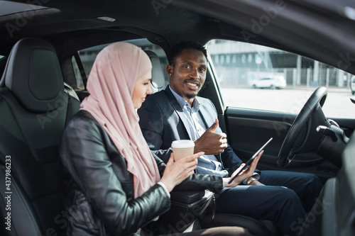 African american businessman in suit and muslim woman in hijab using modern smartphone for remote work while sitting together inside luxury car. Concept of people, gadgets and vehicle. © sofiko14