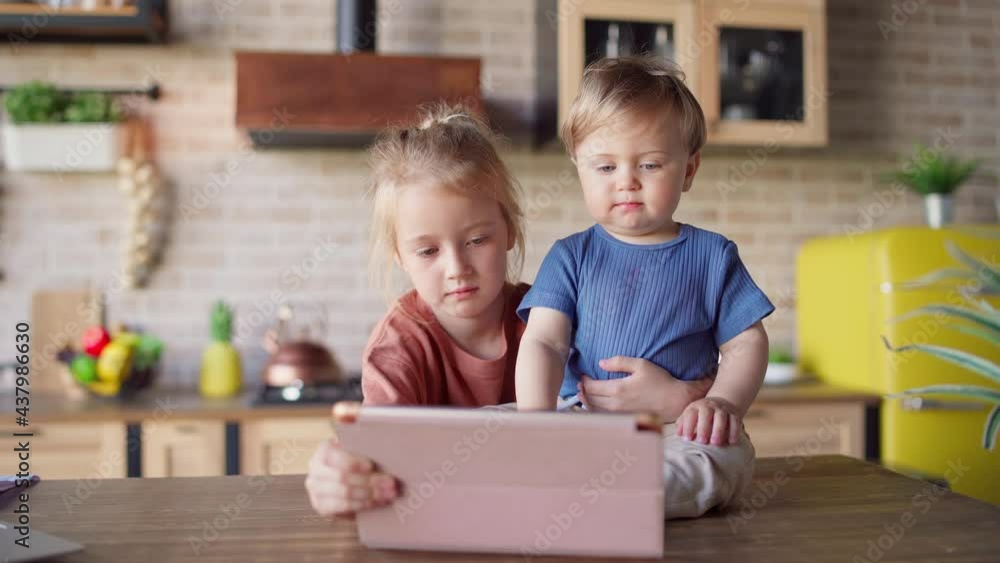 Little girl teaching baby brother how to use tablet computer and play games. Baby boy sitting on kitchen table embraced by loving sister. Siblings playing games on tablet pc together at home Little girl teaching baby brother how to use tablet computer and play games. Baby boy sitting on kitchen table embraced by loving sister. Siblings playing games on tablet pc together at home