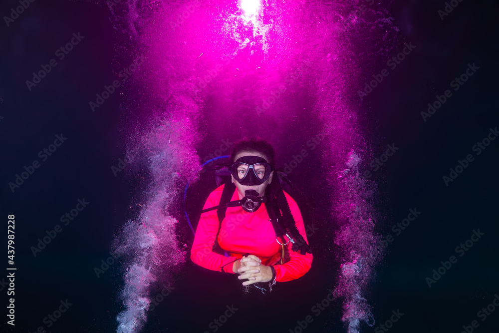 A female scuba diver relaxes into a night dive as explosions of pink bubbles decorate the water around her