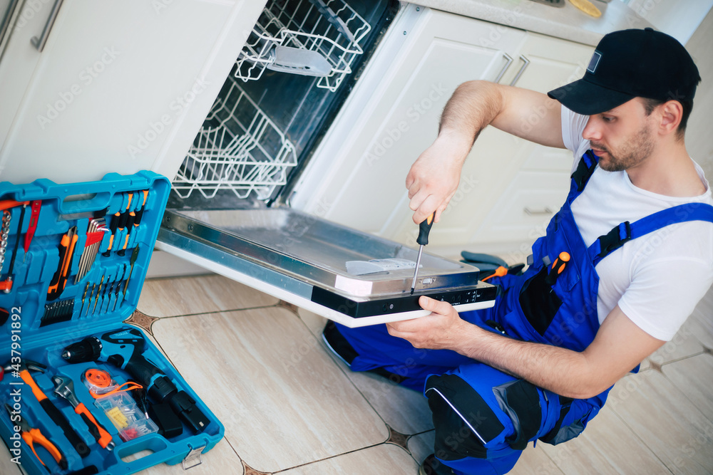 Young modern serviceman in worker suit during the repairing of the dishwasher on the domestic kitchen.