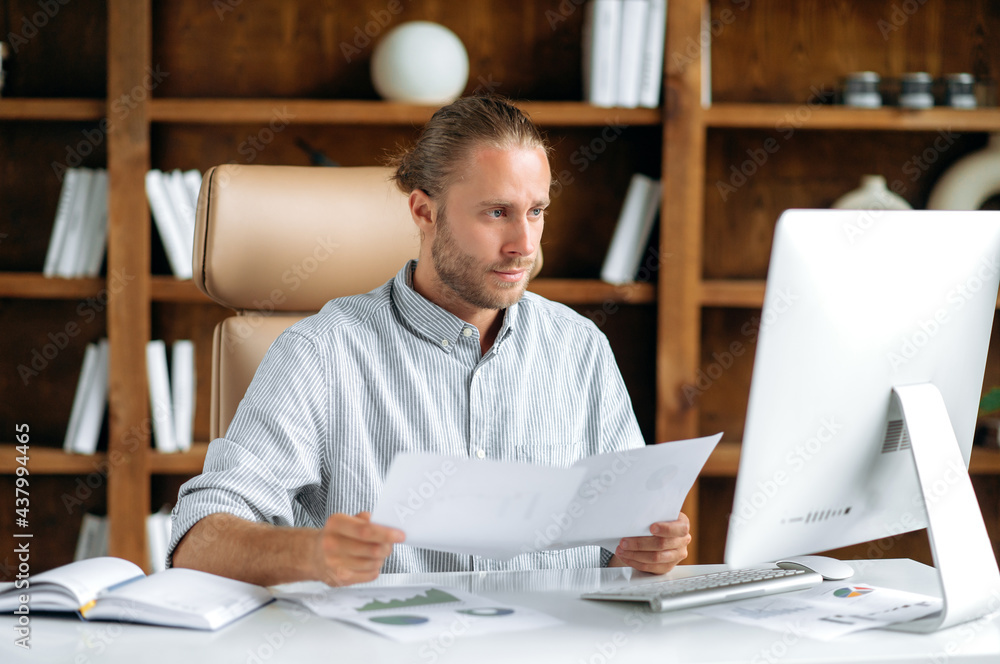 Smart focused young adult caucasian man, sits at the work desk in office, in stylish clothes, uses a computer, looks through documents and charts, thinks over financial strategy, working concentrated Smart focused young adult caucasian man, sits at the work desk in office, in stylish clothes, uses a computer, looks through documents and charts, thinks over financial strategy, working concentrated