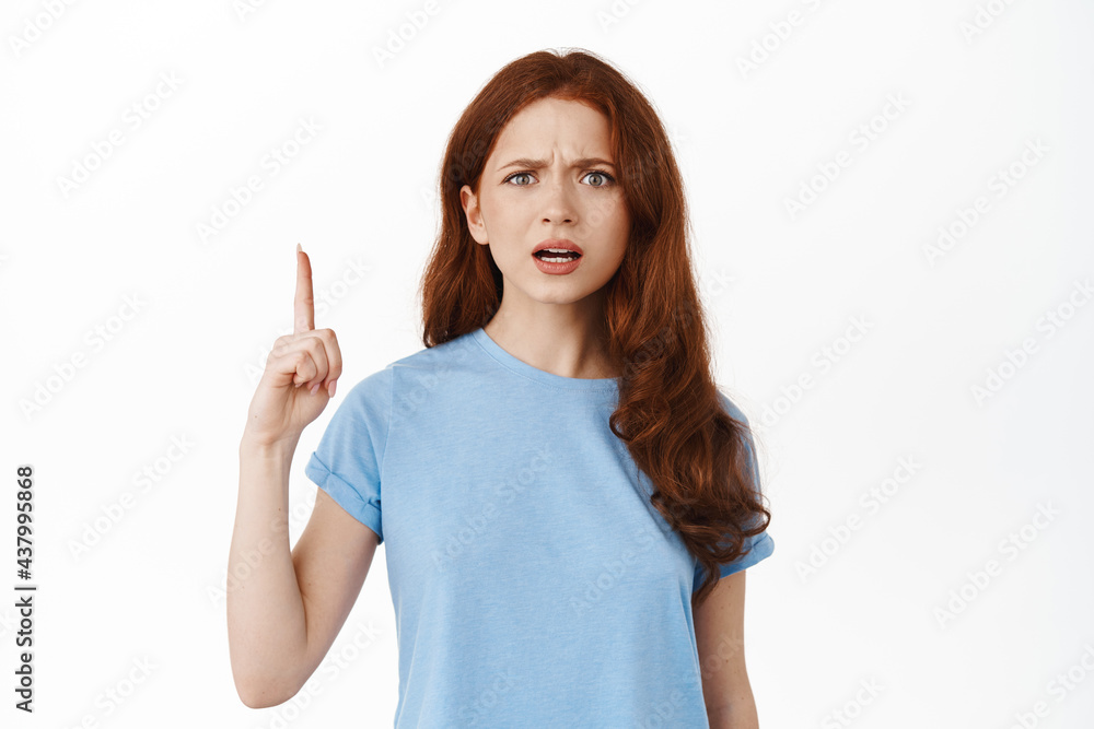 Frustrated and confused redhead woman frowning, pointing finger up at something strange and disappointing, standing in t-shirt against white background Frustrated and confused redhead woman frowning, pointing finger up at something strange and disappointing, standing in t-shirt against white background