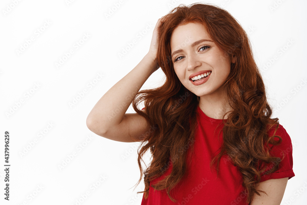Portrait of beautiful natural redhead girl with curly hair, touching head and smiling happy. Candid woman with white smile, wearing red t-shirt, standing against white background Portrait of beautiful natural redhead girl with curly hair, touching head and smiling happy. Candid woman with white smile, wearing red t-shirt, standing against white background