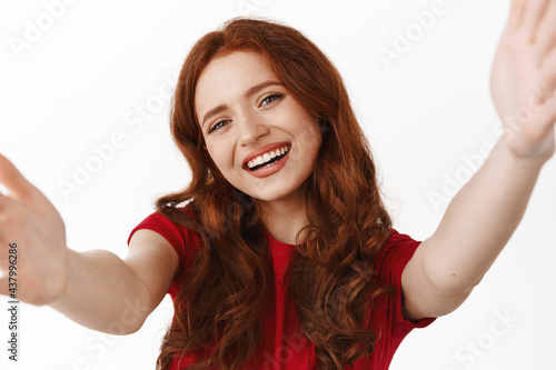 Close up of beautiful redhead curly girl, holding device or tablet, smiling at camera of gadget, video chat or taking selfie, record video, standing in red t-shirt against white background