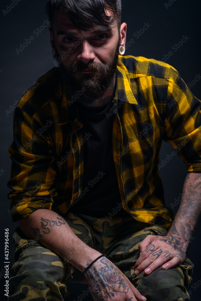 Fototapeta premium Tattooed man with a beard wearing checkered shirt is posing seated on a chair in a studio