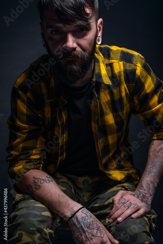 Tattooed man with a beard wearing checkered shirt is posing seated on a chair in a studio
