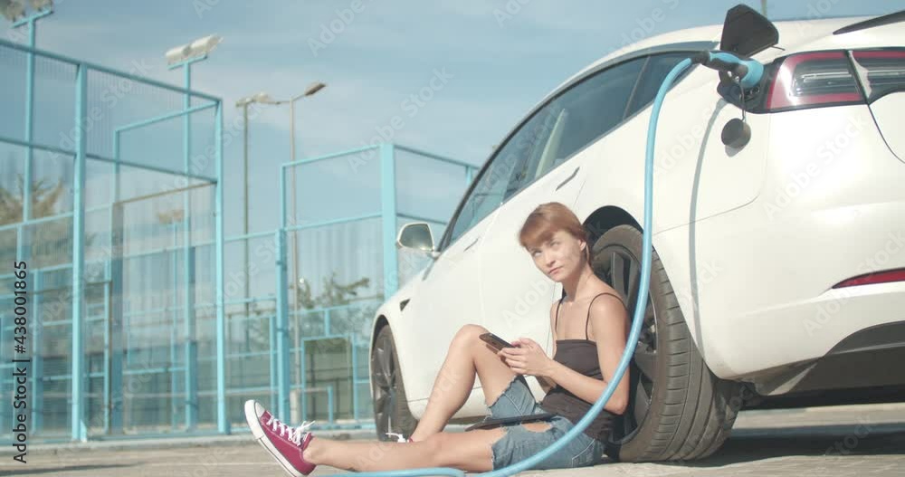 Girl waiting on the ground while her electric car is charging. Taking selfy photo with phone camera Girl waiting on the ground while her electric car is charging. Taking selfy photo with phone camera
