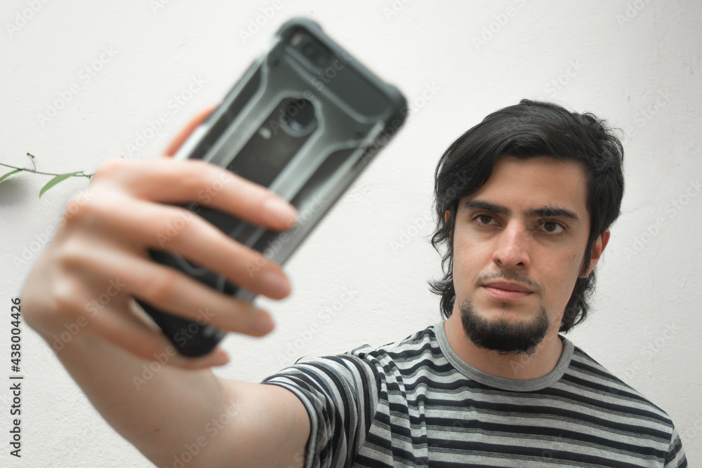 Fototapeta premium Portrait of a young man with beard holding a phone on his hand taking a selfie with serious or handsome expression. Model wearing a black and white striped t-shirt.