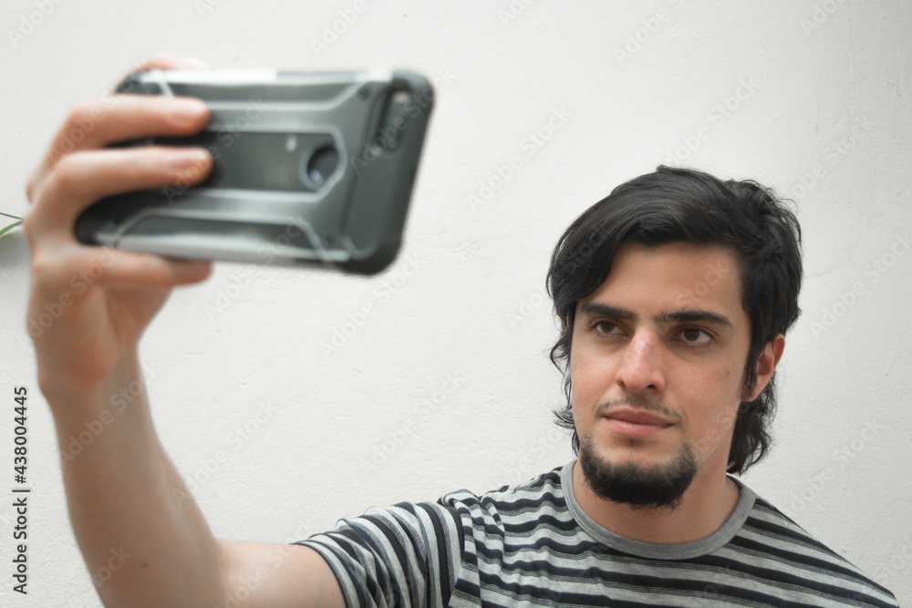 Portrait of a young man with beard holding a phone on his hand taking a selfie with serious or handsome expression. Model wearing a black and white striped t-shirt. Portrait of a young man with beard holding a phone on his hand taking a selfie with serious or handsome expression. Model wearing a black and white striped t-shirt.
