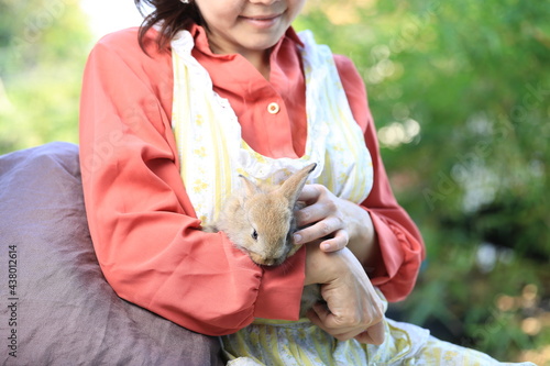 Adorable young rabbit and woman sit together outdoor. Owner care little cute bunny on her laps. Woman wear longsleeves pink redish shirt. photo