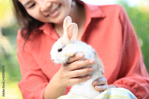 Adorable young rabbit and woman sit together outdoor. Owner care little cute bunny on her laps. Woman wear longsleeves pink redish shirt.