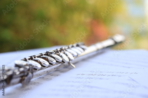 Flute, woodwind brass instrument in classical orchestra. Silver modern flute on white sheet music note for education and performance. Song composer on score sheet with green bokeh nature.