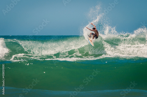 Professional surfer riding a wave and jumping on it with arms raised, at the beach of Praia Brava in Brazil on a sunny day with a blue sky and beautiful aquamarine water.