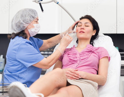 Experienced woman cosmetologist in a protective mask  working in the clinic  introduces a mesotherapy injection into the face of a female patient