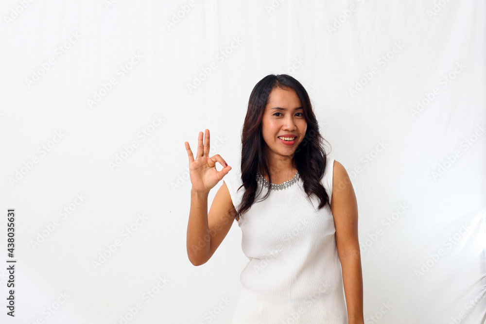 Portrait of a cheerful asian girl dressed in white standing on a white background and doing OK sign with finger and looking at a camera Portrait of a cheerful asian girl dressed in white standing on a white background and doing OK sign with finger and looking at a camera