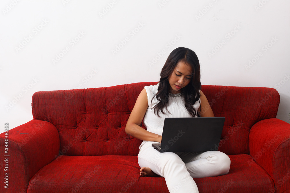 Naklejka premium Asian woman using a laptop while sitting on red sofa. Work from home concept