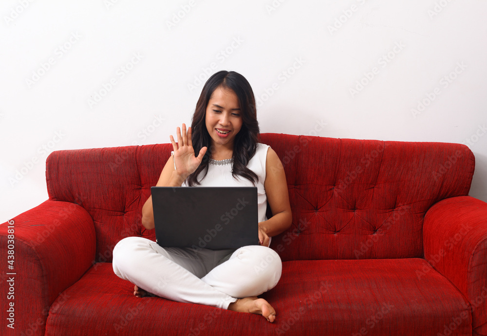 Happy Asian woman using a laptop while sitting on red sofa. Online meeting concept Happy Asian woman using a laptop while sitting on red sofa. Online meeting concept