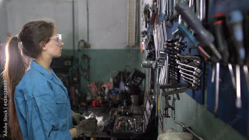 A woman car mechanic in work clothes stands near a stand with wrenches. Selects the required size. Automotive workshop concept. A woman car mechanic in work clothes stands near a stand with wrenches. Selects the required size. Automotive workshop concept.
