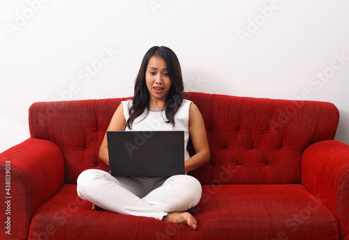 Surprised Asian woman using a laptop while sitting on red sofa.