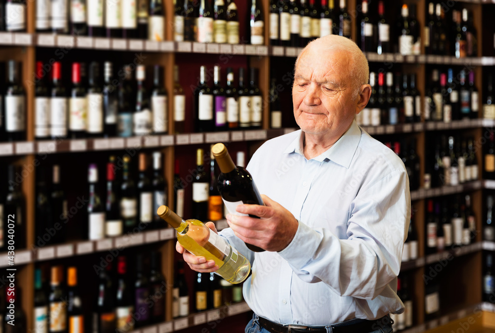 Positive elderly man chooses between red and white wine in a liquor store