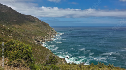 There is sparse vegetation on the slopes of the coastal mountains. The turquoise waves of the ocean beat against the rocks, foaming. Clouds in the blue sky. Cape Town. South Africa